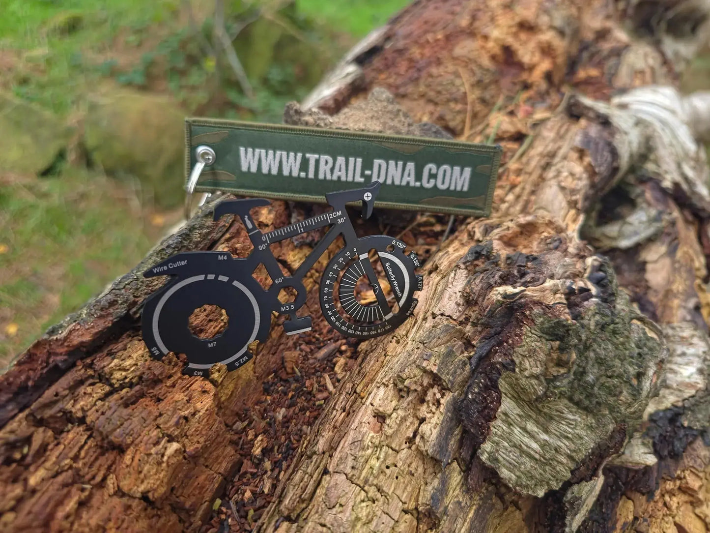 TrailDNA 20-in-1 bike multi-tool with woven keyring (www.trail-dna.com reverse side) resting on a log outdoors