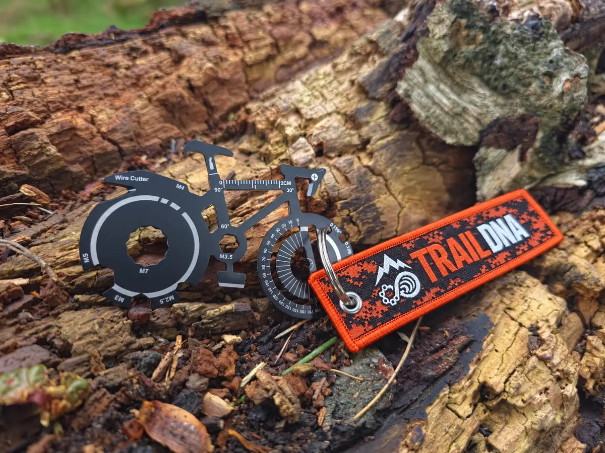 TrailDNA bicycle-shaped multi-tool with orange and black logo keyring on a woodland log.