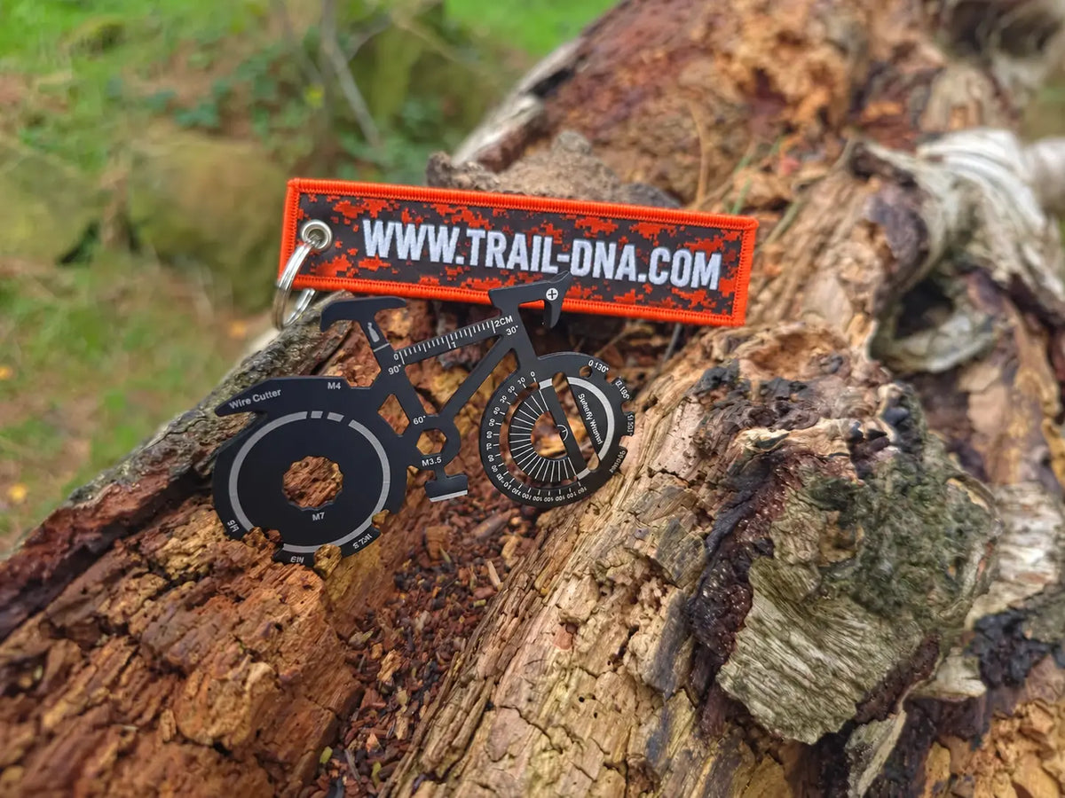 TrailDNA bicycle-shaped multi-tool beside the reverse of the orange keyring, showing WWW.TRAIL-DNA.COM.