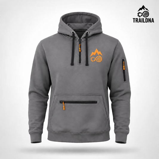 A gray pullover hoodie with black drawstrings, a black half-zip, and orange accents including a mountain and infinity symbol on the chest, a vertical zip pocket on the sleeve, and a horizontal zip pocket on the front. The brand name 'TRAILDNA' is visible in the top right corner.