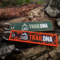Green and orange TrailDNA woven keyrings showing their front logos on a woodland log.