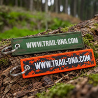 Reverse of green and orange TrailDNA woven keyrings, both showing WWW.TRAIL-DNA.COM, on a woodland log.
