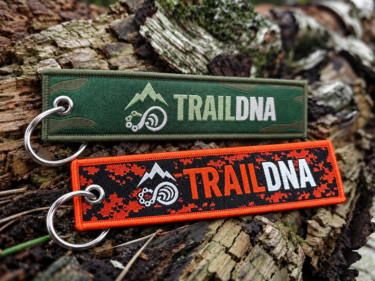 Green and orange TrailDNA woven keyrings with silver split rings resting together on a woodland log.